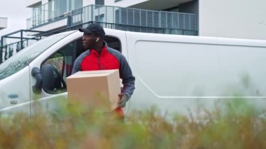 Kırmızı kazaklı ve siyah şapkalı genç bir teslimatçı elinde karton kutuyla beyaz minibüse doğru gülümsüyor. - Evet. Yüksek kalite 4k görüntü