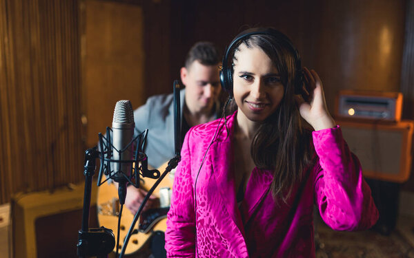 Medium closeup indoor portrait of young caucasian singer woman with headphones on, using recording microphone and looking at camera. Bassist in the background. High quality photo