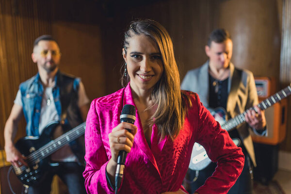 Woman lead singer looks at camera and smiles. Stylish female vocalist standing in front of two guitarists. Performance and music. High quality photo