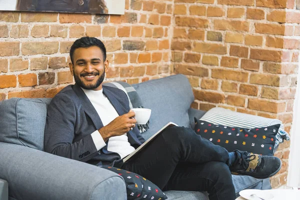 Smiling elegant Indian man relaxing in waiting area of a barber shop. Mid adult man in suit sitting on grey sofa with a cup of tea or coffee. High quality photo