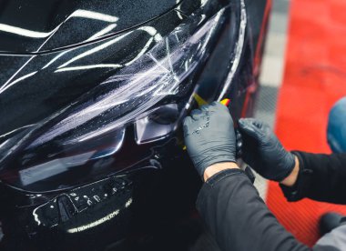İki el koruyucu siyah lastik eldivenlerle arabanın farlarını karartmak için filmle kaplıyor. Kapalı mekanda yakın çekim. Araba tamiri. Yüksek kalite fotoğraf