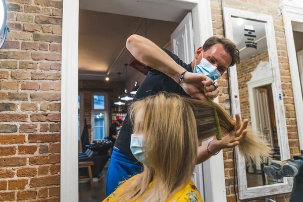 Professional happy male hairdresser with mask styling his clients blonde hair with comb in the professional hair salon. Medium shot. High quality photo