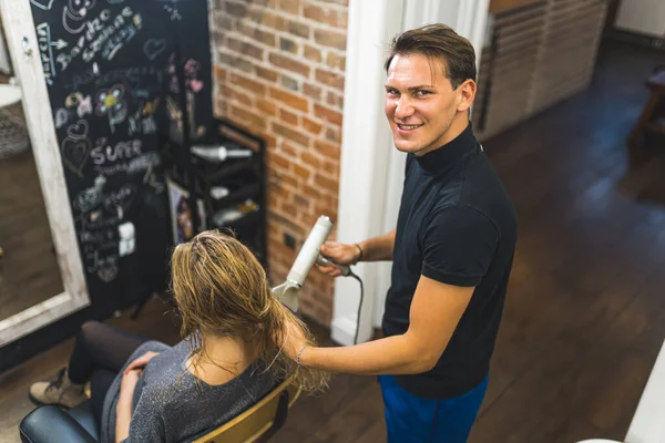 Professional male hairdresser drying and styling his female clients blonde hair with dryer in the professional hair salon. The male hairdresser is looking into the camera. . High quality photo