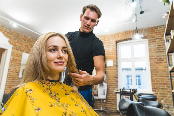 Professional male hairdresser styling his female clients blonde hair with comb and scissors in the professional hair salon. Medium close-up shot. High quality photo