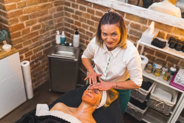 Front view of female beautician putting a beauty mask on her happy blonde clients face. Female beautician looking into the camera. High quality photo
