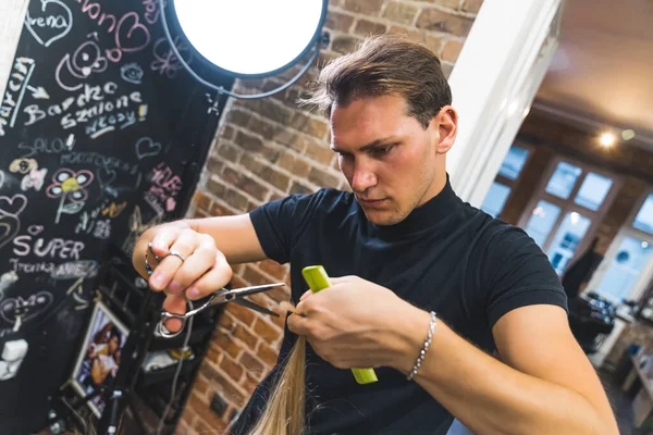 Professional focused male hairdresser styling his clients blonde hair with comb and scissors in the professional hair salon. Medium shot. High quality photo