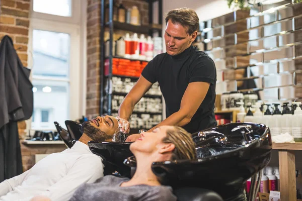 Happy indian male client having his hair washed with shampoo by hairdresser next to blonde female customer in a professional hair salon. High quality photo