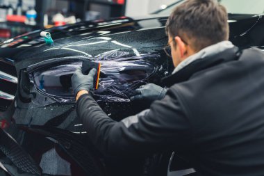 Siyah arabanın yanına çömelmiş bir adamın arka plan görüntüsü. Garajdaki farlara PPF boya koruma filmi uygulamak için. Kapalı alanda yatay çekim. Yüksek kalite fotoğraf