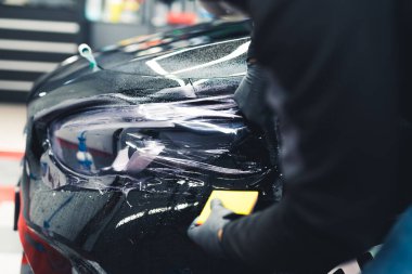 Garajda siyah bir arabanın arkasına PPF boya koruması uygulayan eldiven takan bir adama yaklaş. Araba tamiri. Kapalı alanda yatay çekim. Yüksek kalite fotoğraf