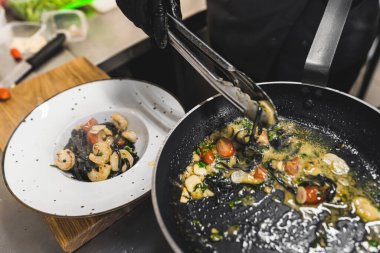 Şef bir yemek hazırlıyor. Beyaz tabağa karides ve siyah tagliolini makarna yerleştirmek için yemek maşası kullanan birinin üst görüntüsü. Yüksek kalite fotoğraf