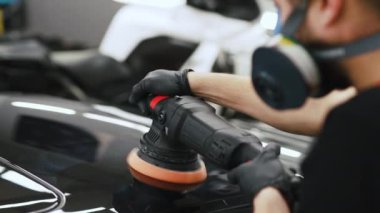 Toz maskeli beyaz bir adam araba cilalamak için cilalama makinesi kullanıyor. Madium shot 'ı. Arka planda bulanık siyah beyaz bir motosiklet görüyoruz. Yüksek kalite 4k görüntü