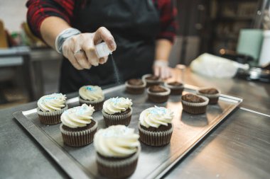Kırmızı ekose gömlekli, siyah önlüklü ve şeffaf eldivenli bir uzman fırın tepsisine mavi serpiştirilmiş kek püskürtüyor. Arka planda dekore edilmemiş kekler var. Arka plan bulanık. Yüksek kalite