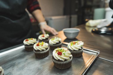 Pişirme tepsisinde beyaz krema ve renkli ışıltılarla süslenmiş altı kek. Mutfakta keklerin yanında duran bir kişi arka planda bulanıklaştı. Yüksek kalite fotoğraf