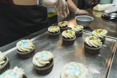 Beyaz kremayla süslenmiş, renkli pullarla süslenmiş altı kekin yan görüntüsü. Ön planda beyaz krema ve mavi süslemelerle süslenmiş bulanık kekler. Bir kişi...