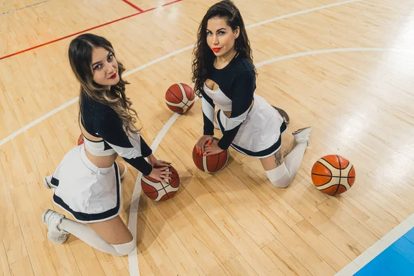 Two vibrant cheerleaders pose in front of the camera, smiling brightly as they hold basketballs on the floor. Dressed in traditional cheer uniforms, complete with mini skirts and knee-high socks.