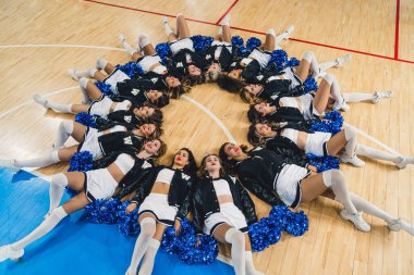 A group of cheerleaders lying on the floor forming a circle with their heads together. 