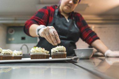 Female baker wearing glove and black apron decorating chocolate frosted cupcakes with sprinkles. Professional baking. Horizontal indoor shot . High quality photo