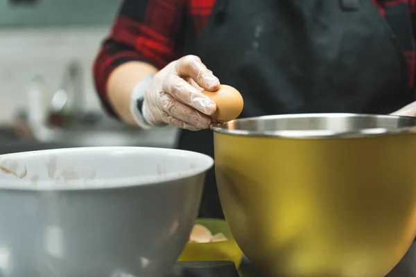 Close-up of female baker wearing gloves and black apron cracking egg into golden bowl to prepare cupcake batter. Professional baking process. Horizontal shot. High quality photo
