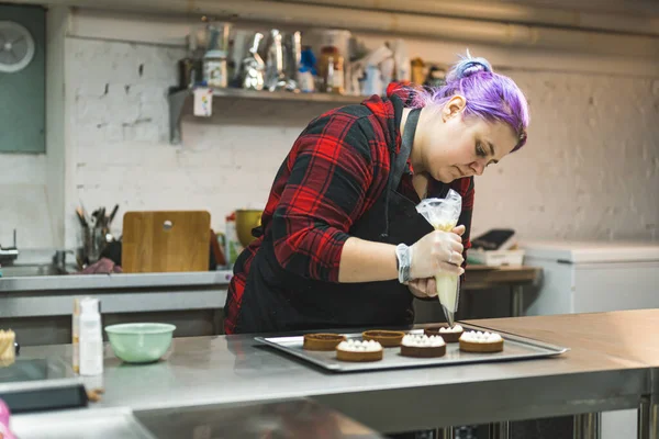Female professional baker with purple hair piping buttercream frosting onto chocolate dessert tarts. Baking process. Horizontal indoor shot. High quality photo