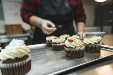 Close-up shot of chocolate cupcakes with white frosting on silver tray being decorated by female baker in black apron. Professional baking. Horizontal indoor shot. High quality photo