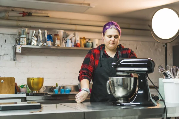 Professional female baker with purple hair wearing black apron making buttercream frosting in stand mixer. Professional kitchen. Baking process. Horizontal indoor shot. High quality photo
