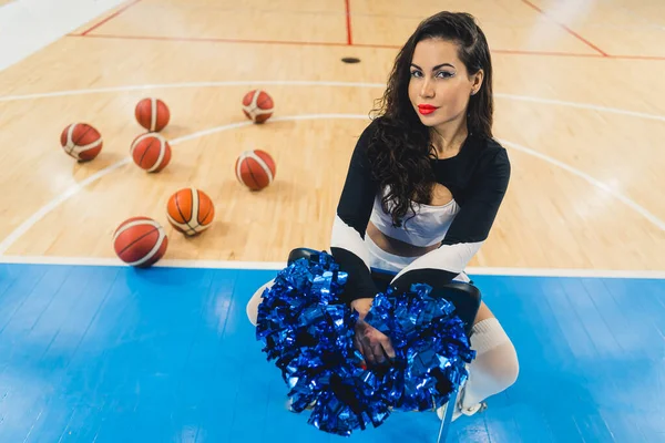High-angle shot of a cheerleader squatting down on a gymnasium floor with blue pom-poms in her hands and basketballs behind her. High quality photo
