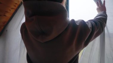 A girl in a gray hoodie open the curtains and we see the view from the window. Outside the window are fields and forests. High quality 4k footage
