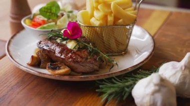 Tasty striploin steak served on mushrooms with French fries. Garlic in the foreground. Wooden table as a background. High quality 4k footage