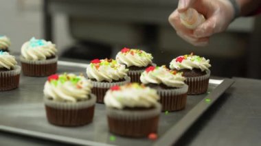 Cupcake decorating process. Close shot of many sweet cupcakes on the foreground while a baker decorating one of them with sprinkles. High quality 4k footage