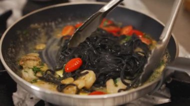 Black tagliolini pasta added to fried shrimps and cherry tomatoes. Restaurant dish. Chef using cooking tongs. . High quality 4k footage