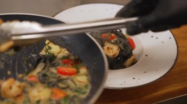 Chef completing a dish. Top view of person using cooking tongs to place black tagliolini pasta with shrimps on white plate. High quality 4k footage