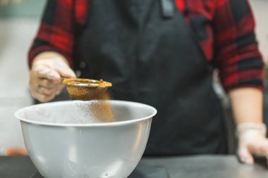 Female profesional baker wearing black apron sifting cocoa powder into white bowl to prepare chocolate cupcake batter. Baking process. Horizontal close-up shot. High quality photo