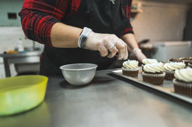 Female professional baker wearing gloves and black apron decorating chocolate frosted cupcakes with blue sprinkles. Horizontal indoor close-up shot. High quality photo
