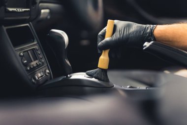 Man hand wearing protective glove, using cleaning brush and removing dust from gear shift knob. Interior of a sports car. Car detailing. High quality photo