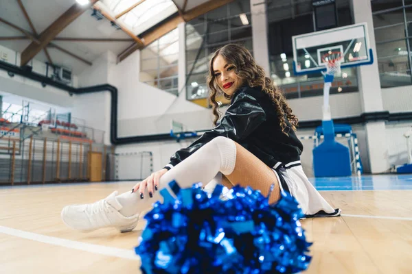 Brunette, curly-haired cheerleader in black and white uniform and jacket sitting on basketball court. Blue shiny pom-pom blurred in the foreground. High quality photo