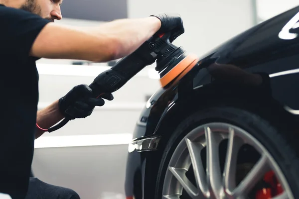 Low angle view of professional car detailing male worker using orbital polisher to polish black car paintwork. Wheel and tyre in the shot. High quality photo