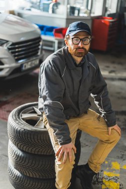 Vertical photo. Medium shot of a bearded caucasian mechanic with glasses sitting on car tyres. Repair shop concept. High quality photo