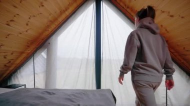 A woman dressed in gray enters an attic room. The ceiling is wooden. The woman opens the curtains. High quality 4k footage