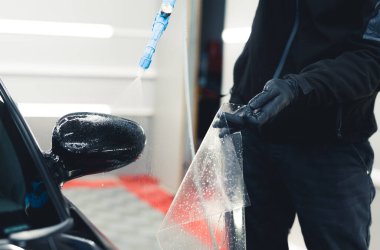 Unrecognisable man wearing black gloves spraying side mirror of car before applying PPF Paint Protection Film. Professional car detailing. Horizontal indoor shot. High quality photo