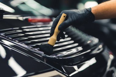 Close-up shot of male hand in black glove wiping brush along detail on trunk of black car. Precision detail work. Car detailing concept. Horizontal indoor shot. High quality photo