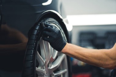Close-up shot of unrecognisable man wearing black glove blackening tires of car using sponge. Professional car detailing in a garage. Horizontal indoor shot. High quality photo
