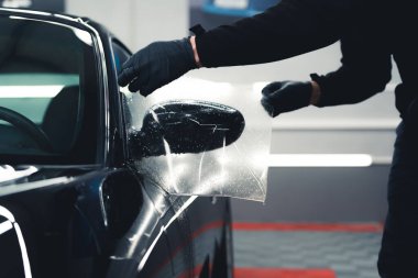 Unrecognisable man wearing black gloves leaning over stretching PPF Paint Protection Film over side car mirror. Professional car detailing process. Horizontal indoor shot. High quality photo
