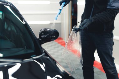 Man wearing work clothes and black gloves preparing side car mirror for PPF Paint Protection Film application. Car detailing process. Horizontal indoor shot . High quality photo