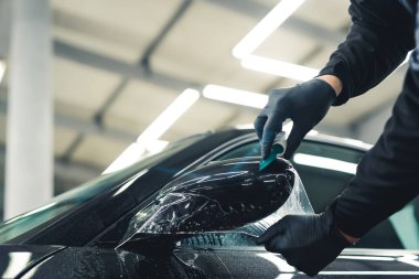 Close-up shot of man wearing black gloves applying PPF Paint Protection Film to side mirror of black car. Car detailing process. Horizontal indoor shot. High quality photo