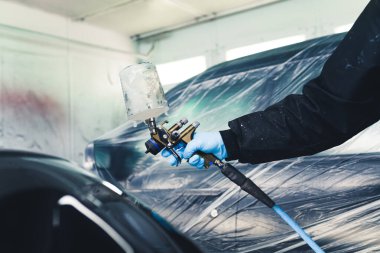 Person wearing protective uniform and blue gloves painting uncovered part of car using airbrush. Car paintwork. Horizontal indoor shot. High quality photo
