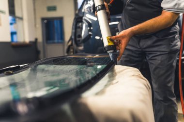 Unrecognisable man applying industrial adhesive to window pane before installing into car. Garage work. Focus on background. Horizontal indoor shot. High quality photo