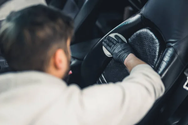 Rear view of man wearing black gloves waterproofing car seat leather upholstery. Protective coating. Professional car detailing process. Blurrred background. Horizontal indoor shot. High quality photo