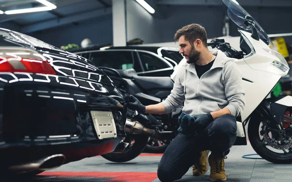 White man kneeling down applying ceramic coating to the back of a black car with motorcycle in the background. Professional car detailing. Garage. Horizontal indoor shot. High quality photo