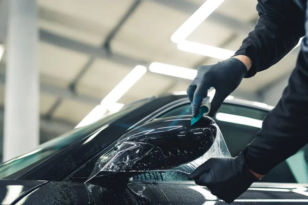 Close-up shot of man wearing black gloves applying PPF Paint Protection Film to side mirror of black car. Car detailing process. Horizontal indoor shot. High quality photo