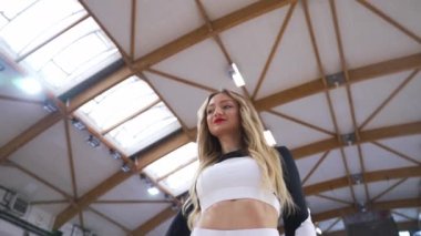 Low-angle shot of a blond cheerleader holding a basketball under her arm and squinting at the camera. Blurred bakground. Sport concept. High quality 4k footage